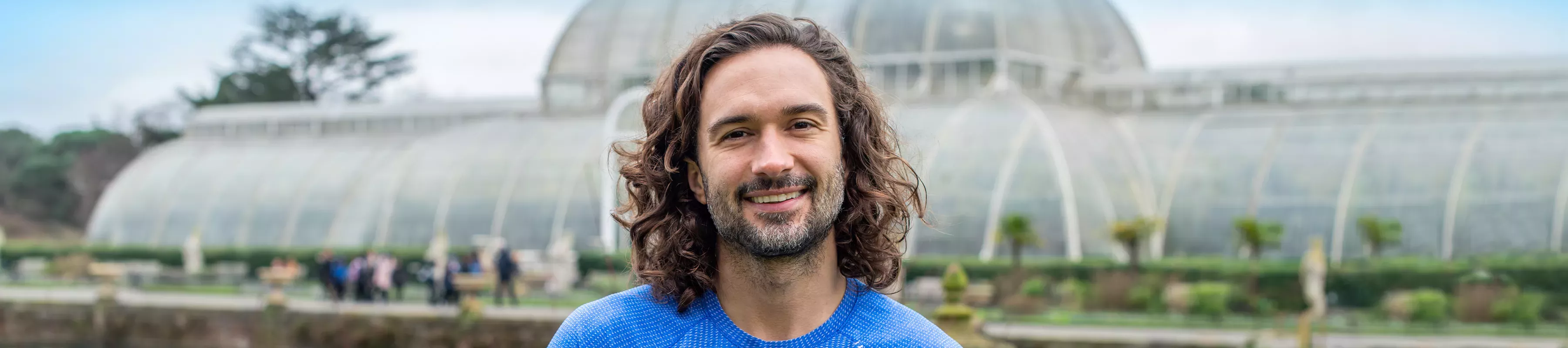 A man with long dark hair, Joe Wicks, stood in front of Kews palm House a large glass building.