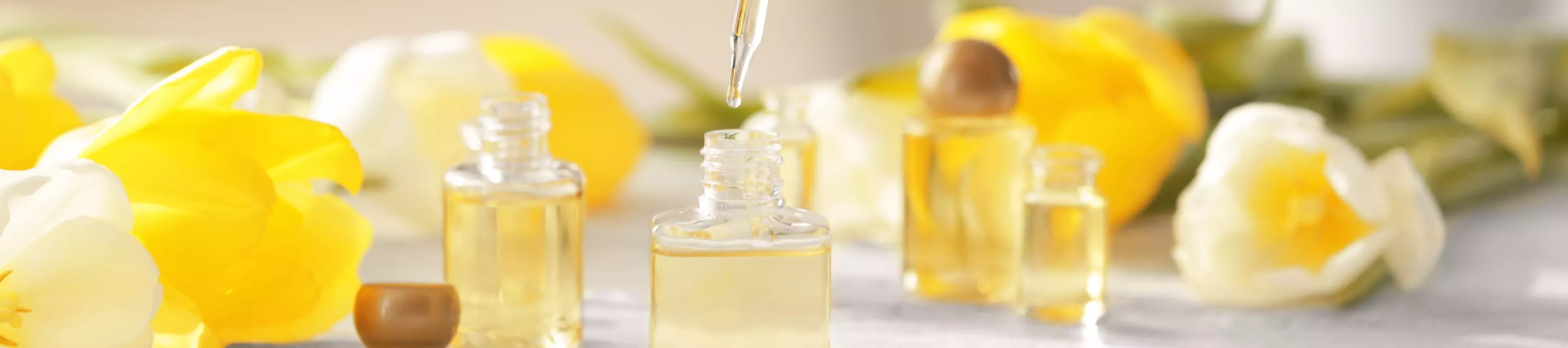 Arrangement of small glass bottles filled with pale yellow oil, with a pipette poised above, surrounded by yellow flowers