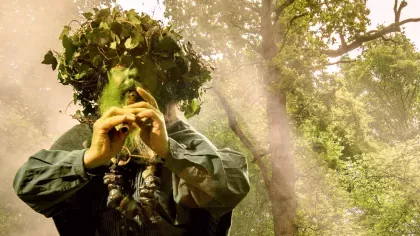 A man in a leafy green costume playing a pair of pipes in a forest