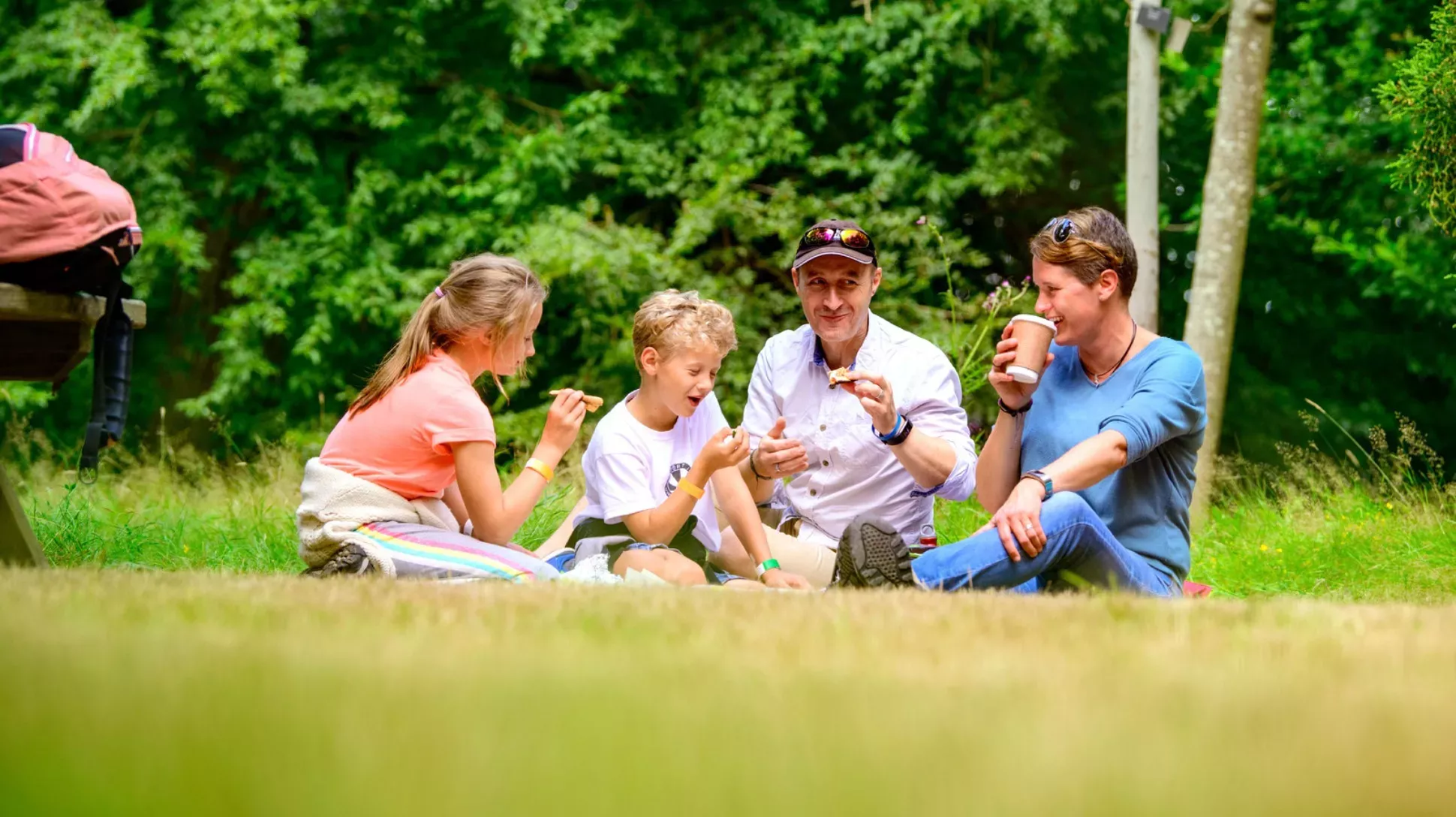 Two parents and two children sit on the grass. They are eating and drinking