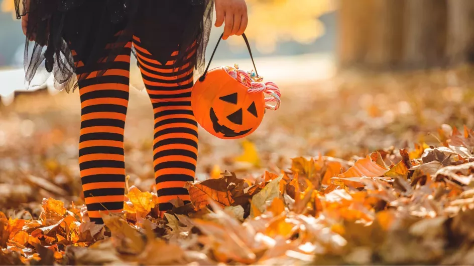 A children in long black and orange socks and holding a plastic pumpkin walking through leaves