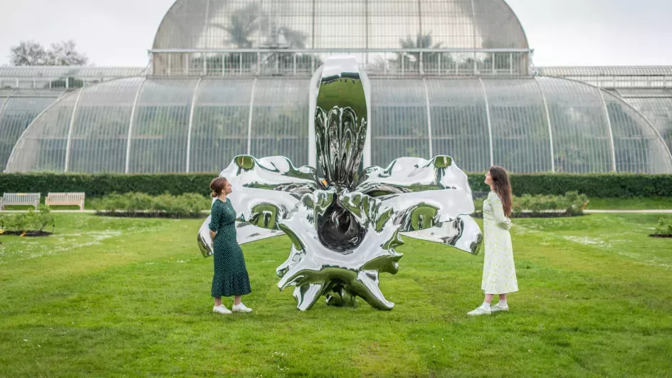 A pair of people look at a shiny metal orchid sculpture in front of the palm house at Kew gardens