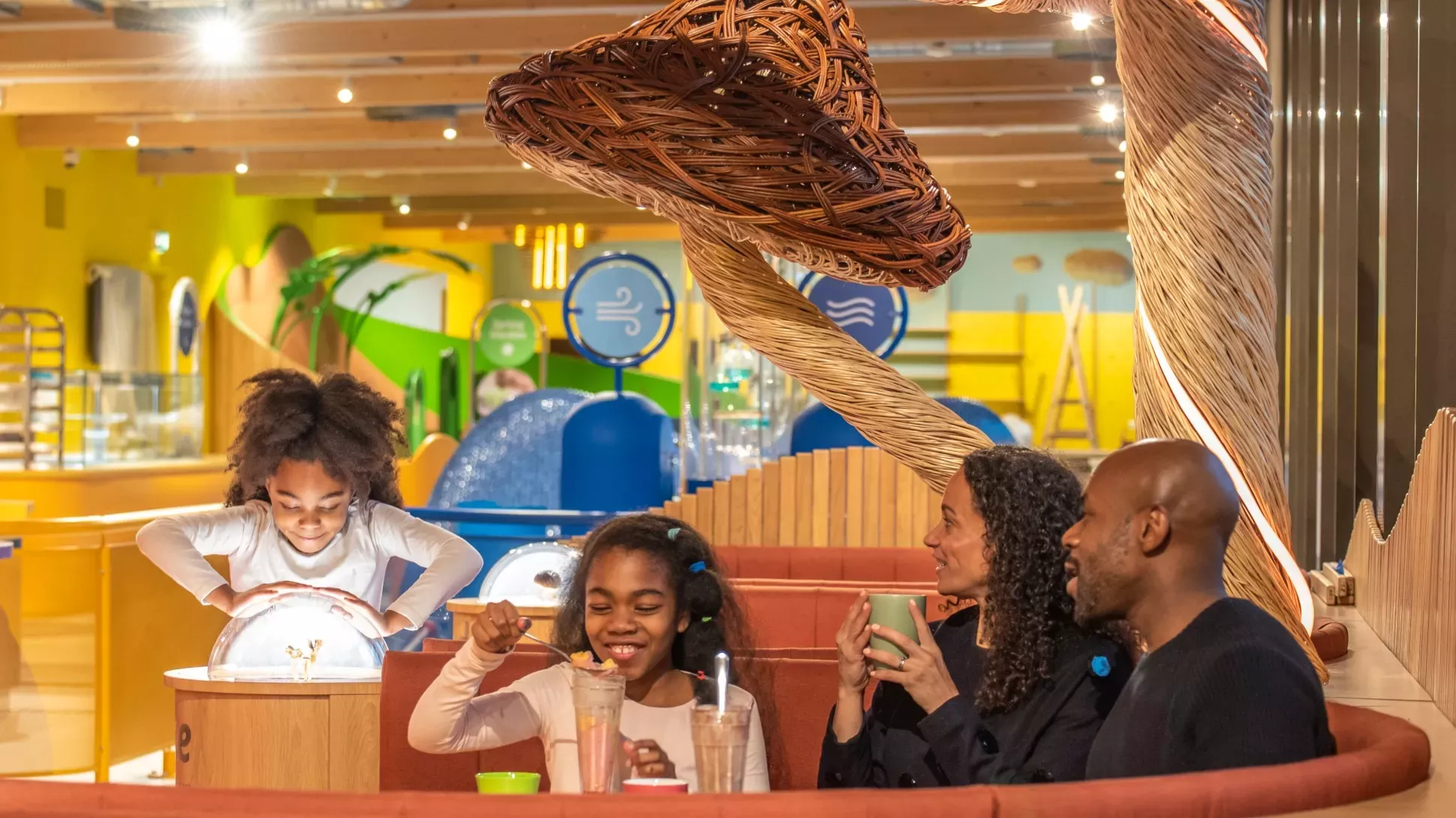 A family of four eats a meal in a colourful restaurant underneath a brown mushroom made from sticks