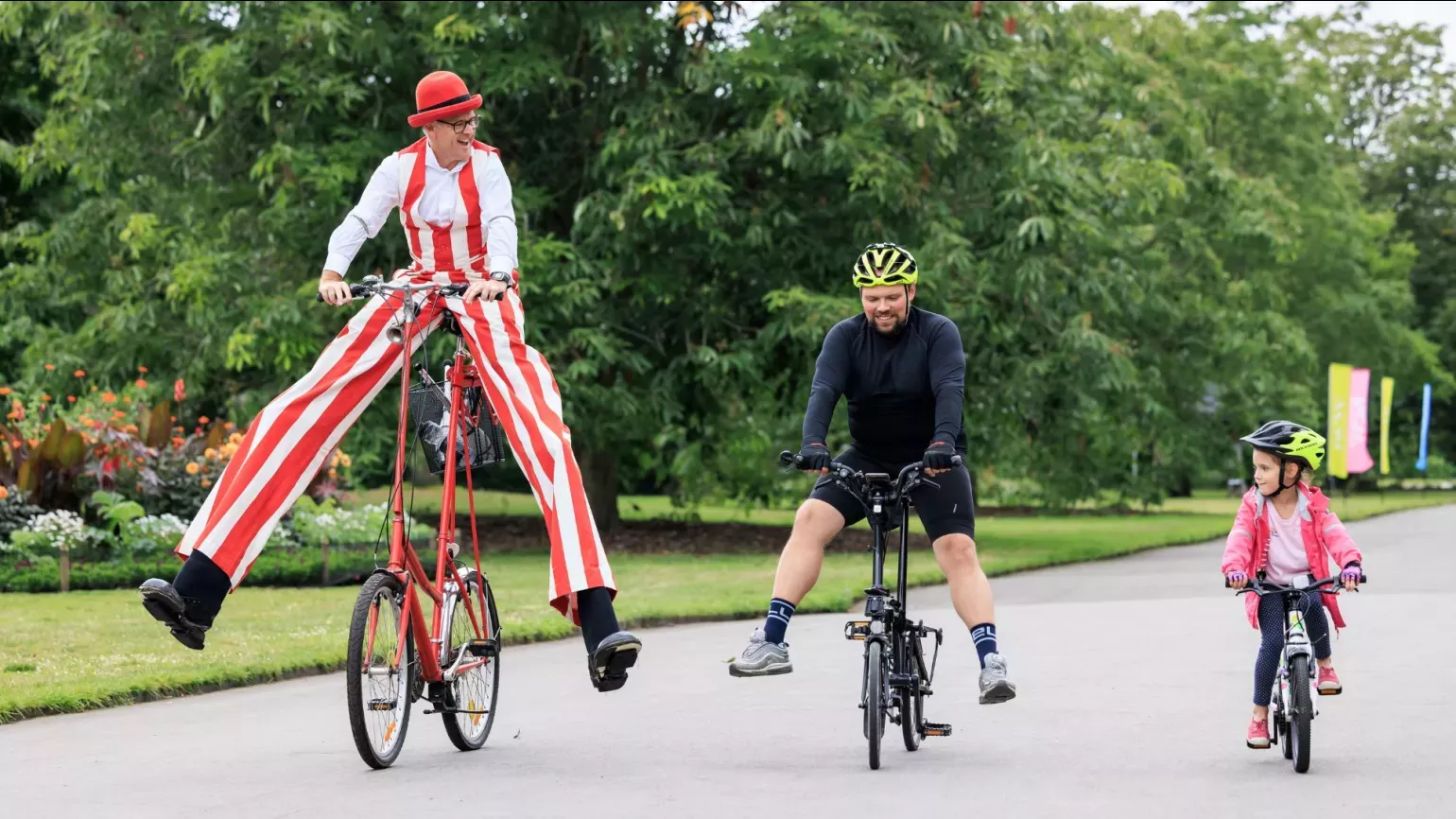 A person in stripy trousers and stilts rides a tall bike next to regular bikers